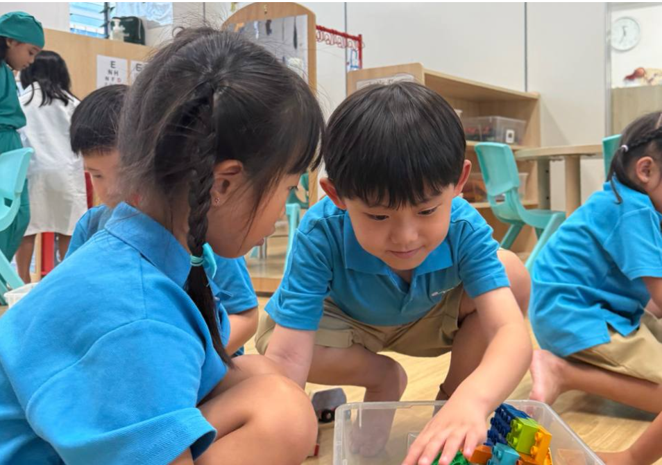 Children in blue uniforms sit on a classroom floor, playing with colorful blocks in a clear bin.